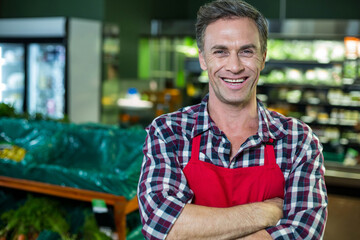 Man standing wearing red apron with arms crossed in produce section among bins, refrigerated cases