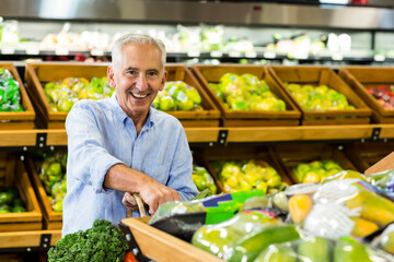Smiling senior man browsing fresh produce in grocery store produce section, holding shopping basket
