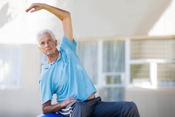 Obraz premium Senior man sitting on blue stability ball stretching left side on outdoor patio wearing blue polo