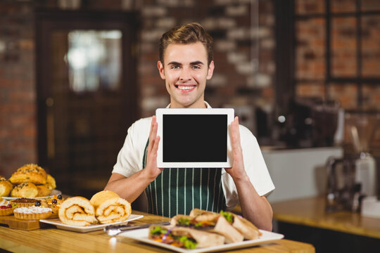 Man holding tablet while wearing striped apron behind wooden counter in café with pastries - Powered by Adobe