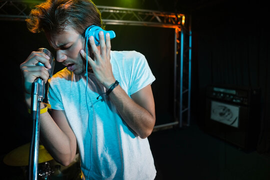 Singer wearing white tee, blue headphones, gripping mic under metal truss on stage, drum kit, amp