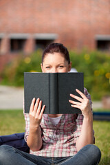 Naklejka premium Black hardcover book resting on grass in campus quad with blurred brick building and green shrubs