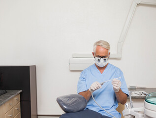 Senior male dentist wearing scrubs and gloves inspecting suction hose at dental chair in exam room