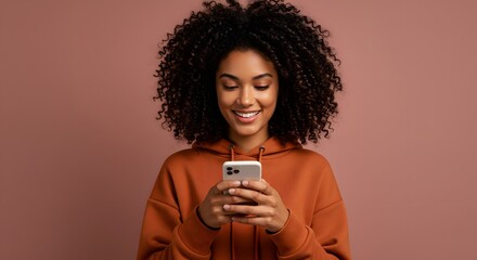 Radiant woman with curly hair in a terracotta hoodie smiling at her smartphone.