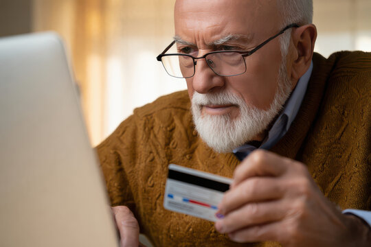 Senior man with glasses holding a credit card looking at a laptop screen with a concerned expression