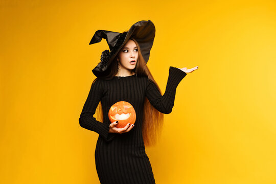 Young woman wearing black dress and hat, holds pumpkin jack o lantern on yellow background. Concept halloween. Girl in witch costume point finger.