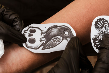 Close-up of a gothic-style bird sitting on a skull tattoo stencil being pressed onto forearm skin...