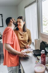 Loving couple sharing a tender moment in the kitchen as they prepare breakfast together. The man kisses the woman on the cheek while she smiles, holding a basket of sliced bread