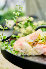 Colorful spring rolls filled with vegetables and shrimp are arranged on a dark, round tray. Lush green plants and delicate flowers surround the dish, creating a lively atmosphere.