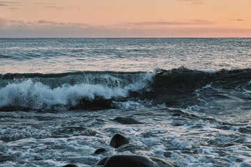 Dynamic ocean wave crashing with white foam against a rocky foreground under a serene evening sky, showcasing the raw power and beauty of the sea at dusk.