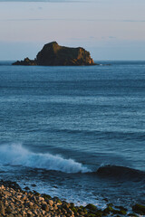 Sunrise over a serene rocky island and pebble beach with breaking waves, offering a tranquil coastal landscape view.