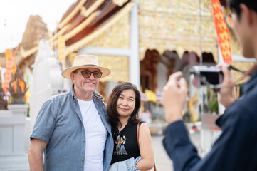 Old white man and asian woman standing and a guide taking picture in front of buddhist golden temple