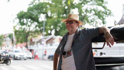 Old white man tourist standing leaning against a tuk tuk or three wheeled taxi after touring a city.
