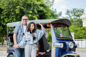 Old white man and asian woman standing posing aside tuk tuk three wheeled taxi after touring a city.