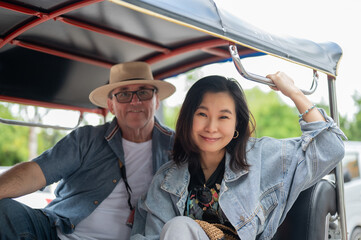 Smiling couple old white man and asian woman sitting in tuk tuk or three wheeled taxi touring a city