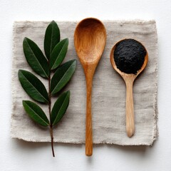 A linen cloth displays two wooden spoons, one holding dark powder, the other empty, and a sprig of vibrant green leaves