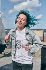 Young woman with blue hair wears denim jacket and white shirt. Standing outdoors with cloudy sky...