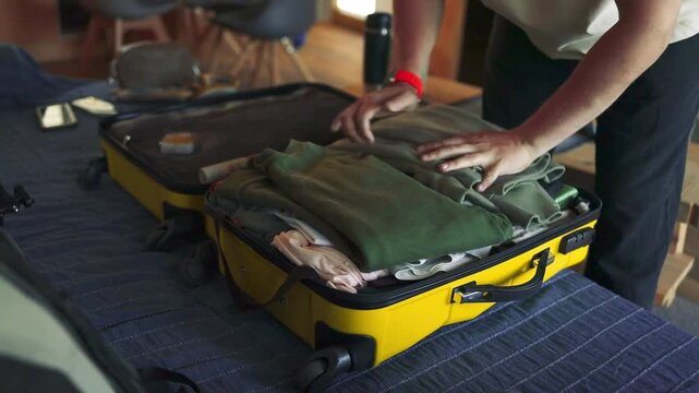 A person packs neatly folded clothes into a yellow hard-shell suitcase on a bed, preparing for a trip. The video captures an indoor travel routine, showing hands placing garments carefully