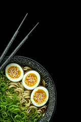 bowl of ramen noodles with boiled eggs, green onions, and sesame seeds with chopsticks isolated on black background