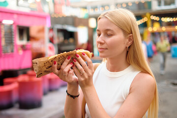 Calm young woman with long blonde hair enjoying hot dog wrapped in decorative paper at vibrant...