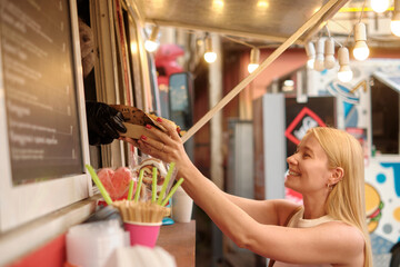 Woman buying hot dog outdoors from food stall, smiling as hand reaches through window, lively...