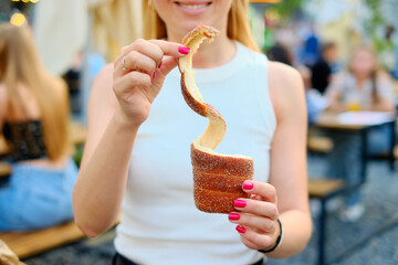 Joyful woman holding a curling cinnamon-sugar coated trdelnik pastry with bright pink nails at...