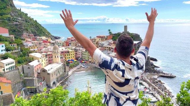 Happy fashion latin backpacker mid adult male tourist raising his arms in celebration while admiring the picturesque Vernazza village in Cinque Terre, Italy