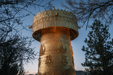 Giant golden prayer wheel at sunset in Shangri-La, Yunnan, a symbol of Tibetan spirituality surrounded by mountains, trees, and traditional Buddhist culture