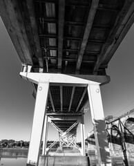 Black and white low-angle view beneath a steel bridge with cross-bracing and support columns