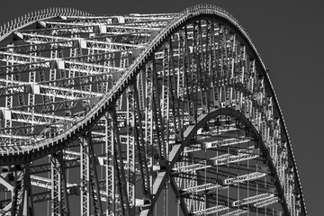 Black and white detailed view of a large steel arch bridge, showing intricate lattice framework and repeating geometric patterns in strong sunlight