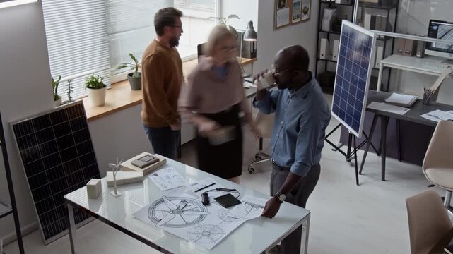 Full surveillance time lapse shot of three multiethnic developers working in office on innovative renewable energy devices, examining wind turbine sketches and solar battery prototypes