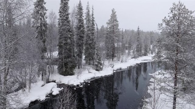 Aerial, slowly flying through snow-covered trees to a river while it's snowing