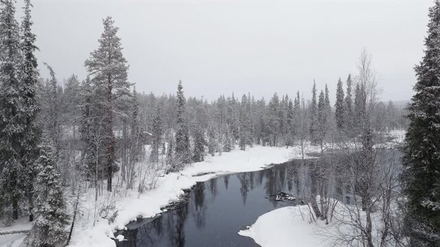 Aerial, slow flight above a river surrounded by snowy forest while it's snowing