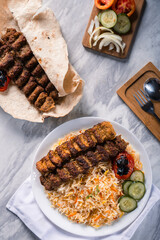 Top view of grilled beef kebab with colorful basmati rice, roasted tomato, cucumber slices, yogurt dip, fresh vegetables, and pita bread on marble surface with wooden cutlery tray beside