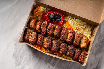 Grilled beef kebab served on colorful basmati rice with roasted tomato and vegetables in a brown takeaway box, placed on a marble surface under natural lighting, showing rich texture and flavor