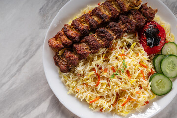 Grilled beef kebab served with colorful basmati rice, slices of fresh cucumber, and a roasted tomato on a white ceramic plate, placed on a marble surface in natural light