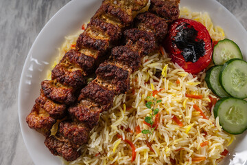 Grilled beef kebab served with colorful basmati rice, slices of fresh cucumber, and a roasted tomato on a white ceramic plate, placed on a marble surface in natural light