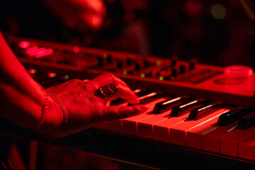Close-up of a person playing a keyboard during a live concert, with red stage lighting reflecting...
