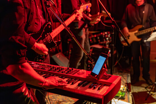 Live music performance with keyboard and electric guitar players on stage, lit by red stage lighting. A smartphone displays lyrics or chords on the keyboard. - Powered by Adobe