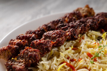 Close-up of grilled beef kebab served with colorful basmati rice and roasted tomato on a white ceramic plate, placed on a light marble surface under soft natural lighting