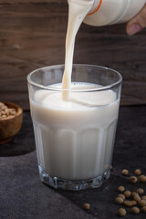 Fresh soy milk being poured into a clear glass on a dark stone surface, with scattered soybeans and a wooden bowl in the background, illustrating natural and healthy plant-based nutrition