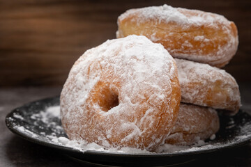 Close-up of stack of sugar-coated donuts on a black plate, with powdered sugar scattered around, placed on dark stone surface and wooden background, highlighting texture and sweetness