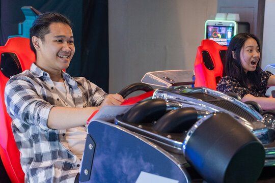 Young Asian couple playing a racing simulator arcade game, sitting in red gaming seats with steering wheels, expressing joy and excitement in a brightly lit entertainment center - Powered by Adobe