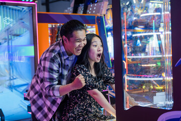 Excited young Asian couple playing an arcade game together in a neon-lit game center, both showing expressive reactions of joy and surprise while looking at the screen in a fun indoor atmosphere