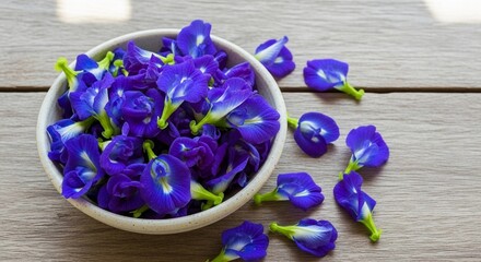 Naklejka premium A bowl filled with vibrant blue butterfly pea flowers on a wooden surface. Some flowers are scattered around the bowl, showcasing their unique shape and color.