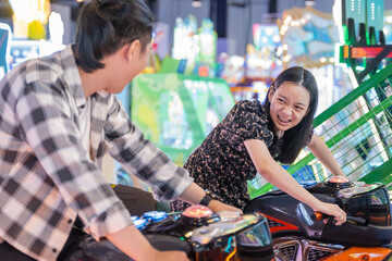 Young Asian couple playing a motorbike racing arcade game, leaning forward with excitement and joy,...