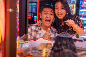 Excited young Asian couple playing an arcade game together, pointing enthusiastically through a...
