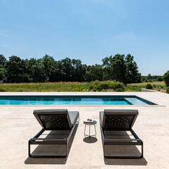 deux transats sur une terrasse au soleil en été devant une piscine à l'eau turquoise vue sur la campagne de l'herbe et une forêt