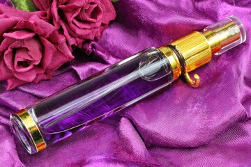 Naklejka premium A cylindrical, clear glass perfume bottle filled with a deep purple liquid, sits atop a draped, purple fabric. Two maroon roses rest in the background