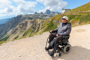 Man with disability using wheelchair enjoys the scenic beauty of Dolomites in Italy. Bright sky serves as backdrop as he navigates the rugged terrain of this stunning mountain range.